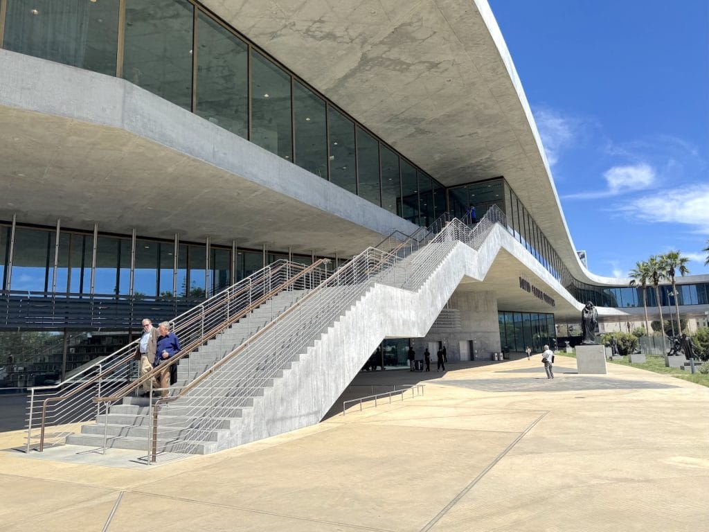 A wide-angle, exterior view of the concrete David Geffen Galleries at LACMA. Modernist stairs ascend toward glass walls under a sweeping, cantilevered roof and blue sky.