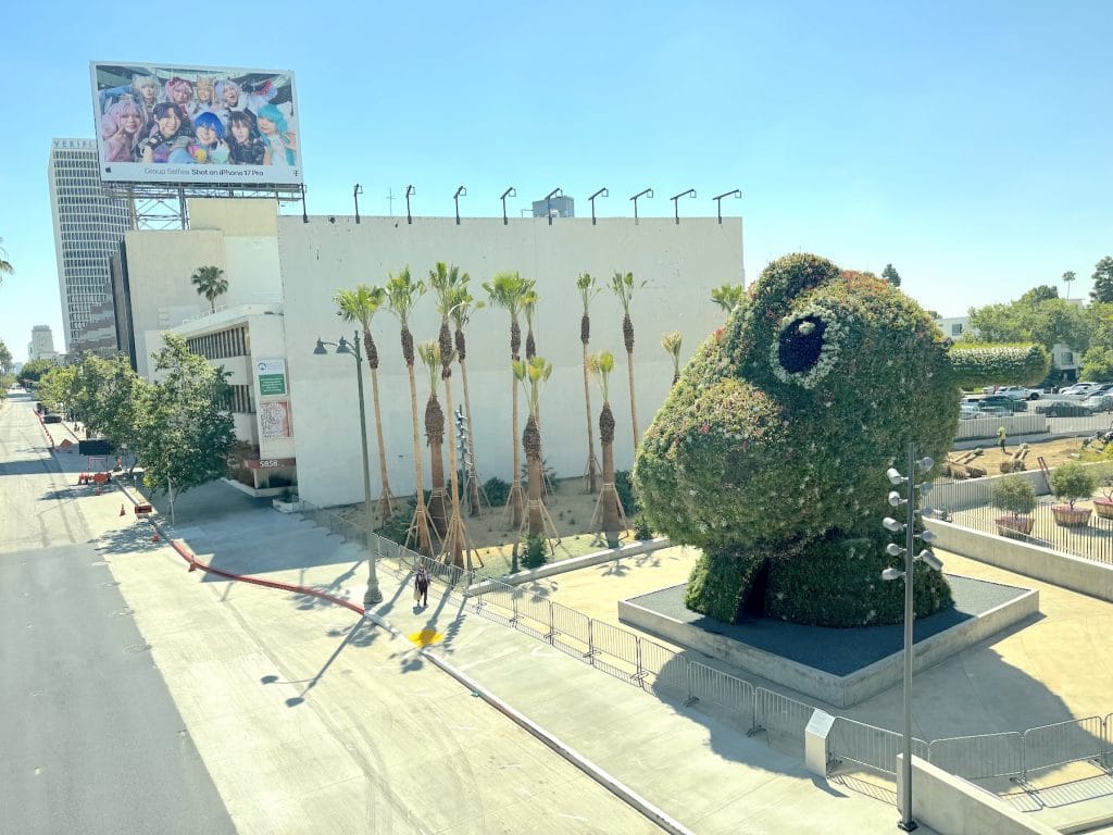 A massive, flowering topiary sculpture of a split rocking horse head stands outdoors near palm trees and a large billboard overlooking a bright city street.