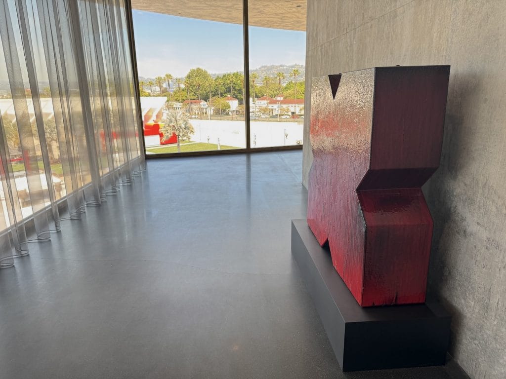 A geometric red-and-black glazed ceramic sculpture stands on a black base against a concrete wall, next to large windows overlooking a bright city landscape.