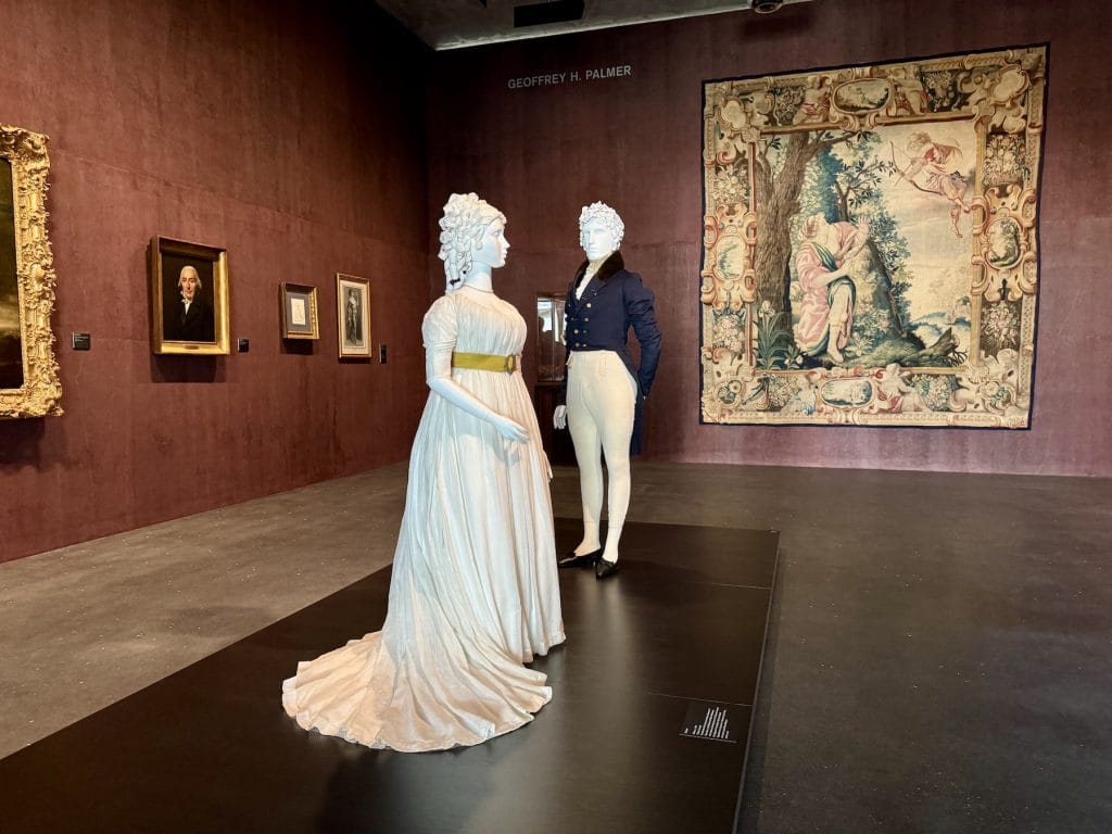 Two mannequins dressed in 19th-century attire stand before a large tapestry in a gallery with deep red walls. Framed portraits hang to the left.