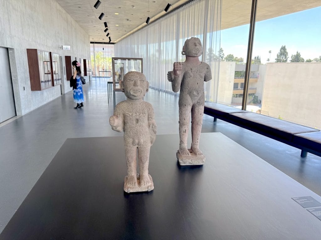 Two ancient stone "Xipe Totec" standard bearer figures stand on a black plinth in a minimalist gallery. Expansive windows with sheer curtains overlook a city.