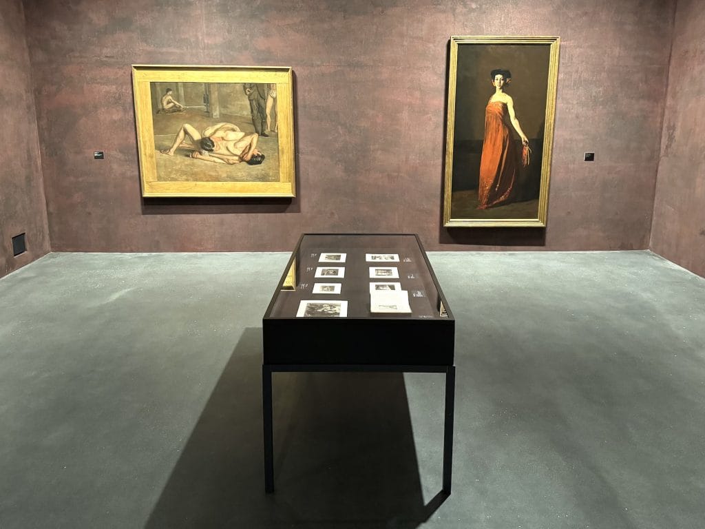 Two oil paintings in gold frames hang on a dark red wall behind a glass display case containing photographs in a quiet museum gallery space.