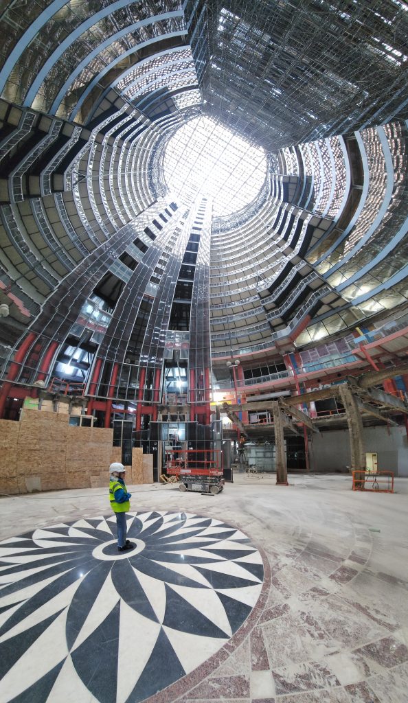Judy Chicago standing at center of Thompson Center atrium, elevator glass wall behind.