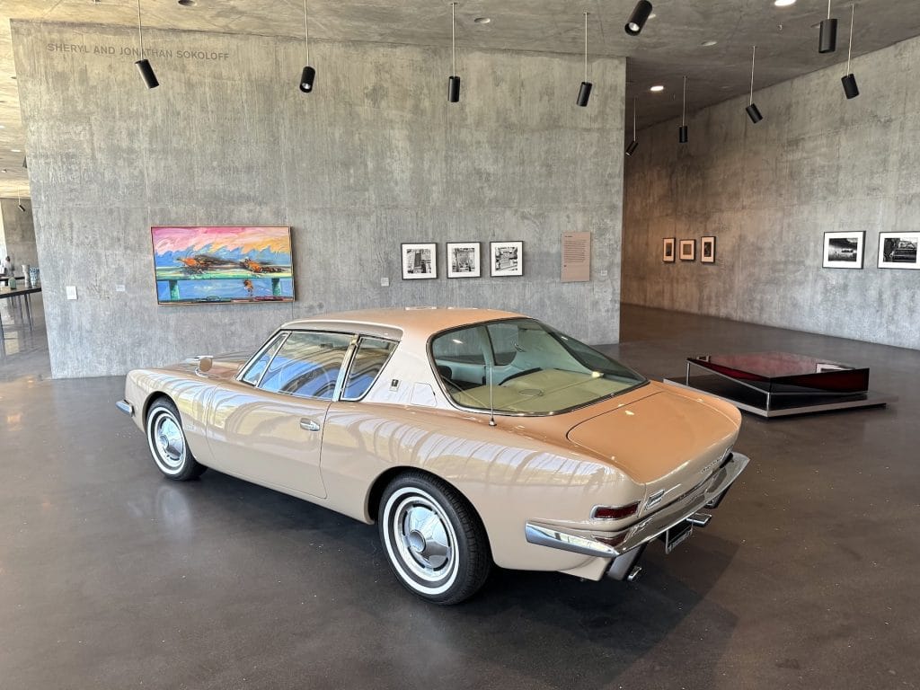 A vintage, tan Studebaker Avanti is displayed in a modern gallery with concrete walls. Colorful paintings and black-and-white photographs hang in the background under spotlights.
