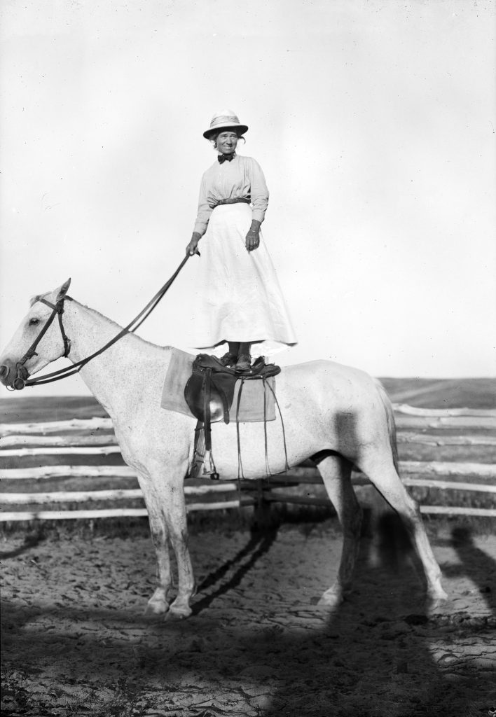 Evelyn Cameron wearing a hat, smiling, holding reins, and standing on her white horse, Jim. [no date]