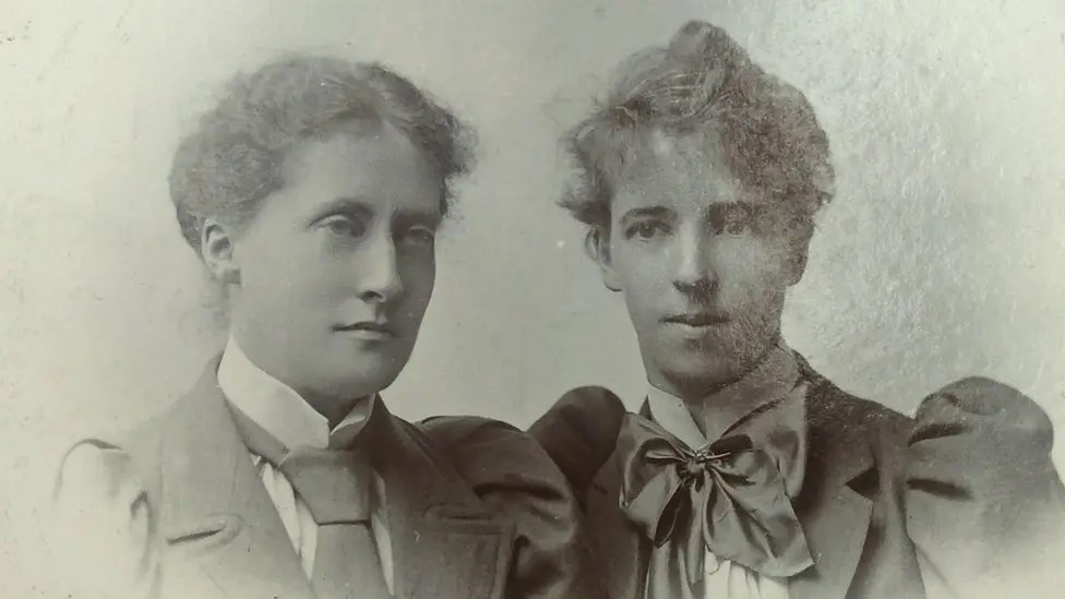 a grainy black-and-white photograph of two women with their hair up and wearing turn of the century suits