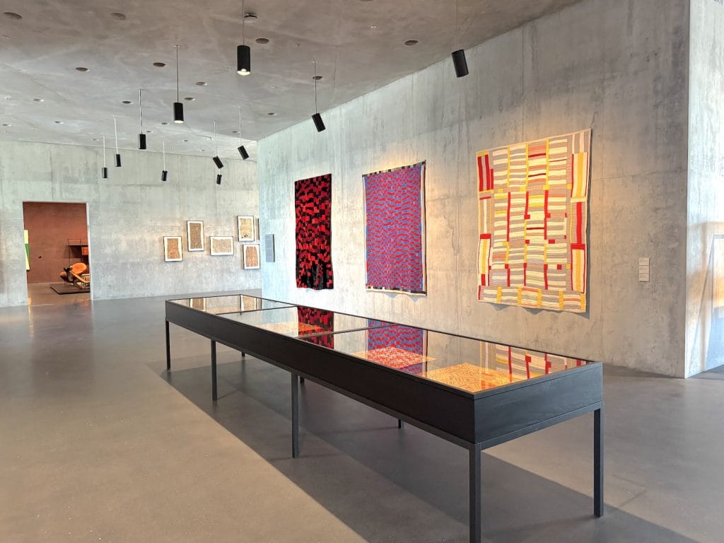 Three vibrant, patterned textile artworks hang on a concrete gallery wall behind a long glass display case containing smaller fabric samples under black pendant lights.
