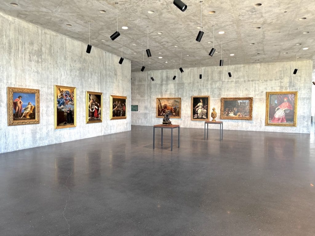 Eight classical oil paintings in ornate gold frames hang on a minimalist concrete wall in a spacious museum gallery illuminated by hanging black track lighting.