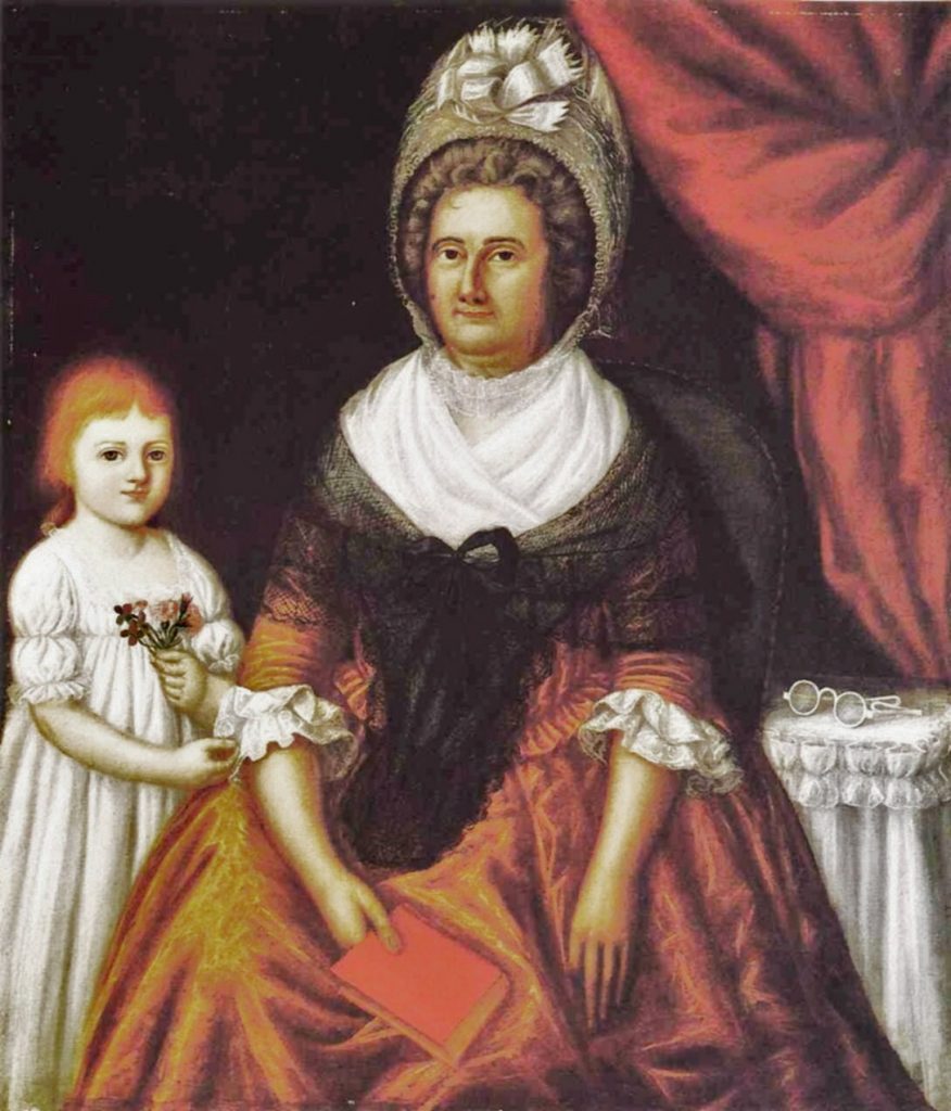 An early American portrait painting by Joshua Johnson of a seated woman and a young child posed indoors against a dark background with a red draped curtain. The woman wears a high white bonnet with a bow, a white kerchief, and a dark shawl over a warm-toned dress with ruffled cuffs. She holds a small red book in her lap and faces forward with a composed expression. Beside her stands a young child in a white dress holding a small sprig of flowers. On a small round table to the right rests a pair of spectacles.