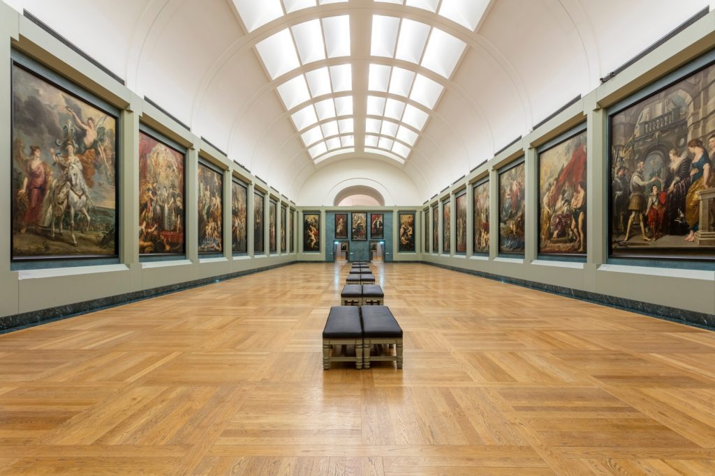 a large museum gallery has paintings lining its green-toned walls and is lit by skylights above