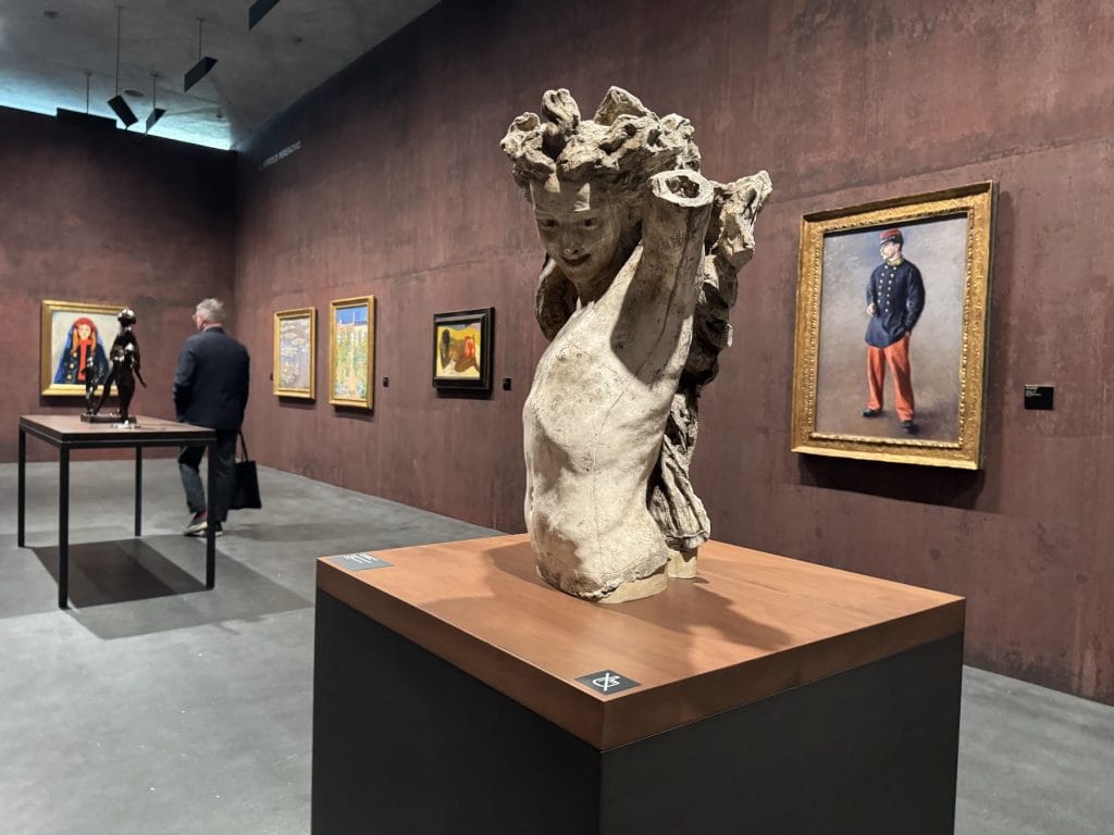A sculpture of a bacchante by Jean-Baptiste Carpeaux stands in a gallery, surrounded by Impressionist paintings on textured reddish walls and a visitor.
