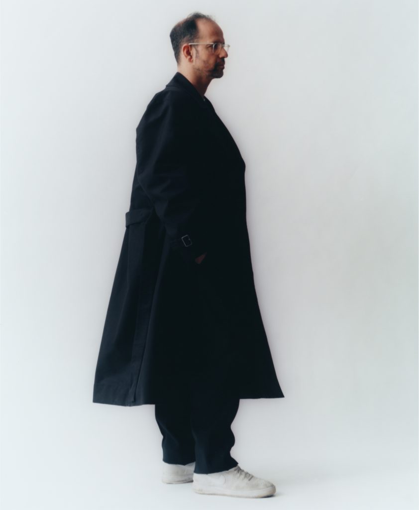 Artist Tino Sehgal standing in profile against a white wall, wearing a long black coat and white sneakers.