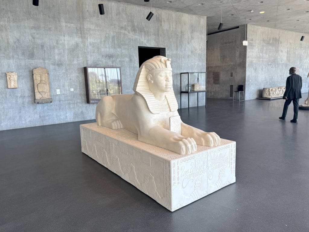 A white sphinx sculpture by Lauren Halsey sits in a concrete gallery. The base features contemporary engravings, blending ancient Egyptian aesthetics with modern community iconography.