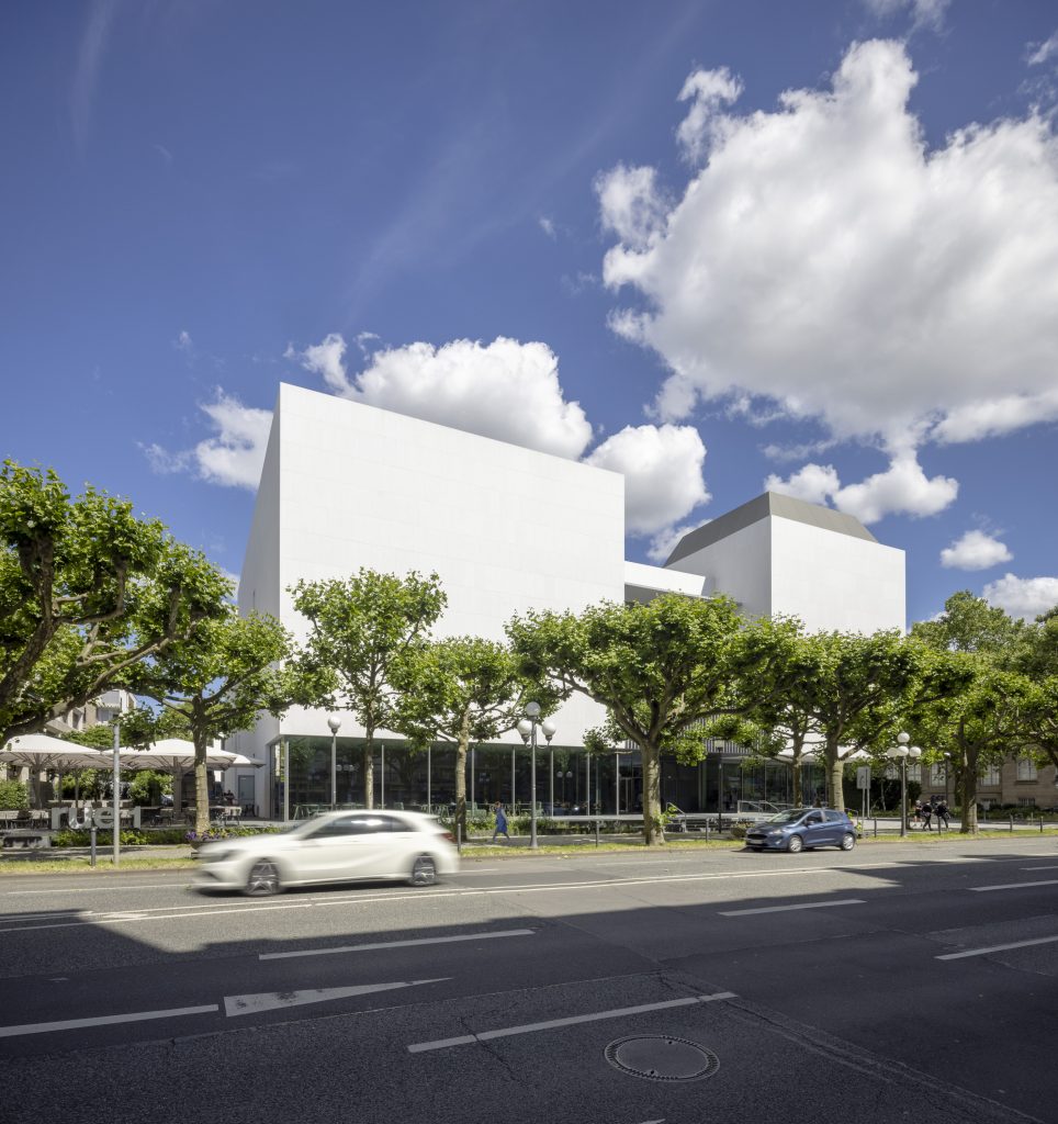 A large white building under the blue sky. There are green trees in front of the building and a white car on the road in front of the building.