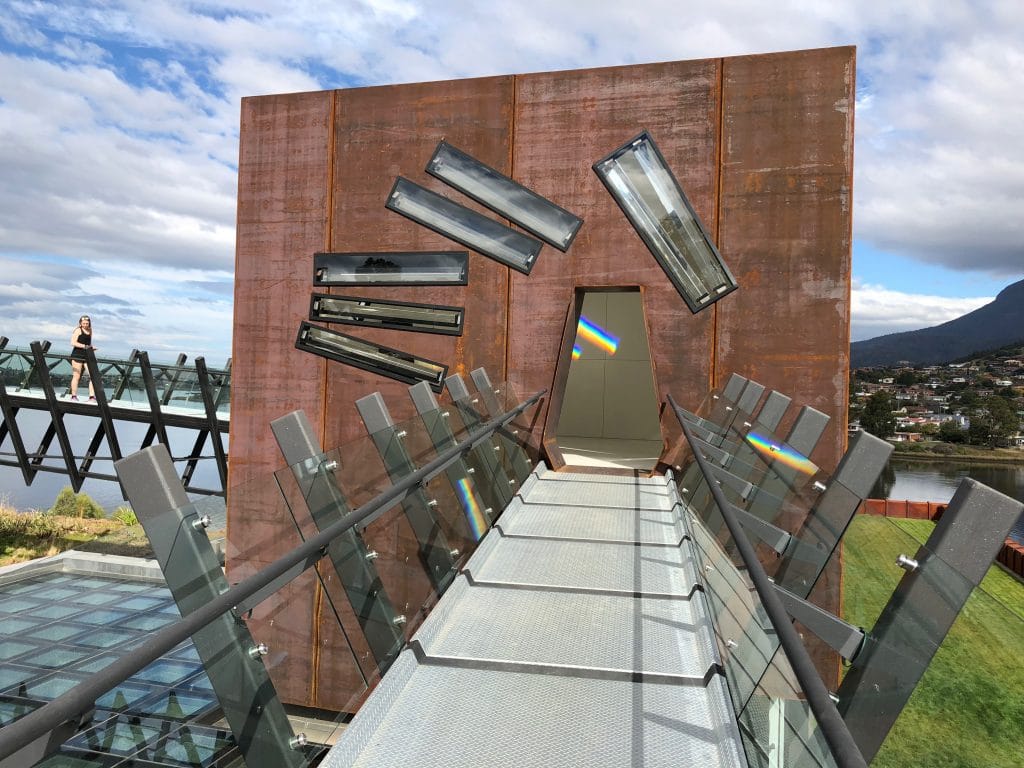 Rust-colored steel building with angled windows at the end of a glass walkway, with rainbow light reflections visible.