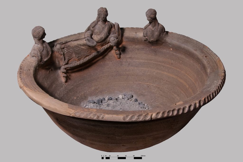 A photograph of a small plain brown ceramic vessel fetauring three small carvings of women around its rim, with just a little bit of burnt material near the bottom of its bowl