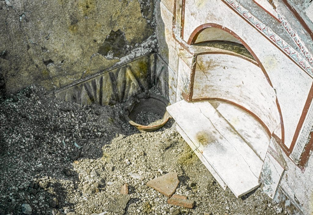 A photograph of a small clay vessel on the ground at Pompeii with a white structure vaguely resembling an old fireplace to its right