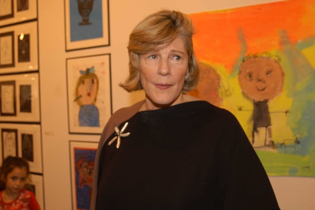 A photo of an older white woman with short blonde hair wearing a dark top with a brooch, standing in front of colorful children's artwork.