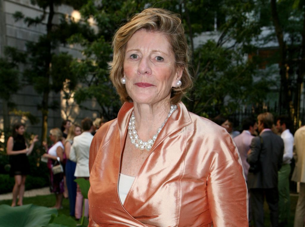 A photo of an older white woman with short blonde hair wearing a shiny peach jacket and pearl necklace, standing outdoors at a social gathering.