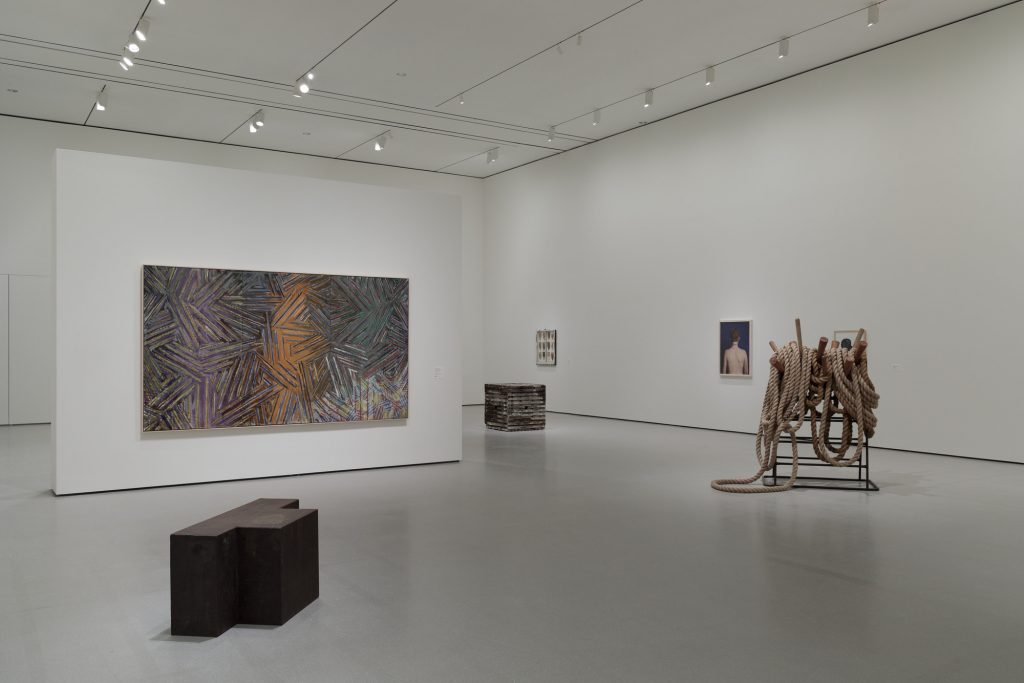 A photo of a modern art gallery with white walls and polished gray floors, displaying large-scale contemporary works including an abstract painting with radiating lines in earthy tones, a cube-like sculpture, a portrait of a nude back, and a rope installation draped over a metal frame.