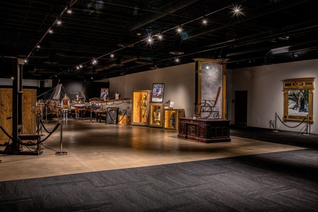 A photo of a large museum gallery with dim lighting and spotlights illuminating historical exhibits. Displays include a small wooden boat, suits of armor, and artifacts arranged behind ropes and in cases, with a large framed painting and decorative columns on the walls.