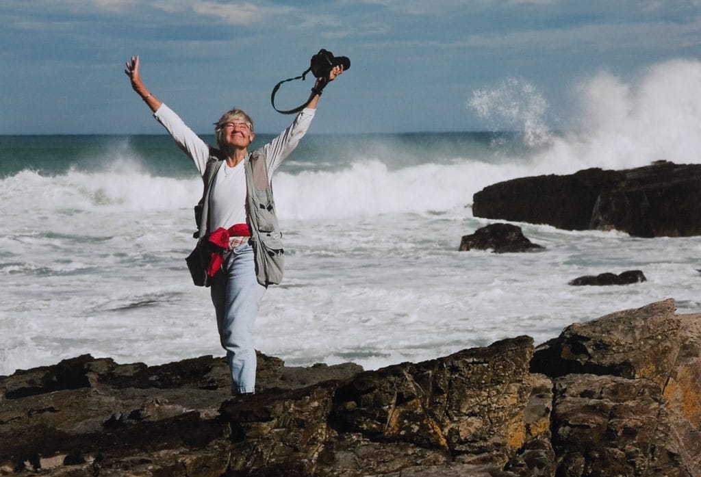 Color photo of a woman standing on rocky shoreline with arms raised, holding a camera, as waves crash behind her.