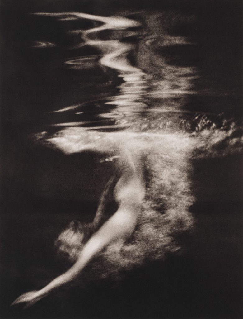 A soft-focus, black-and-white photograph of a female figure submerged underwater, her body arcing diagonally as light ripples across the surface above her, creating abstract, shimmering distortions.