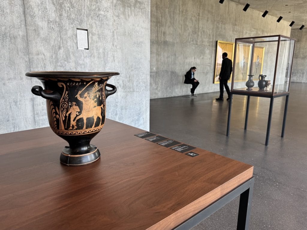An ancient Greek red-figure bell-krater rests on a wooden pedestal in a concrete gallery. In the blurred background, visitors walk past additional glass display cases.
