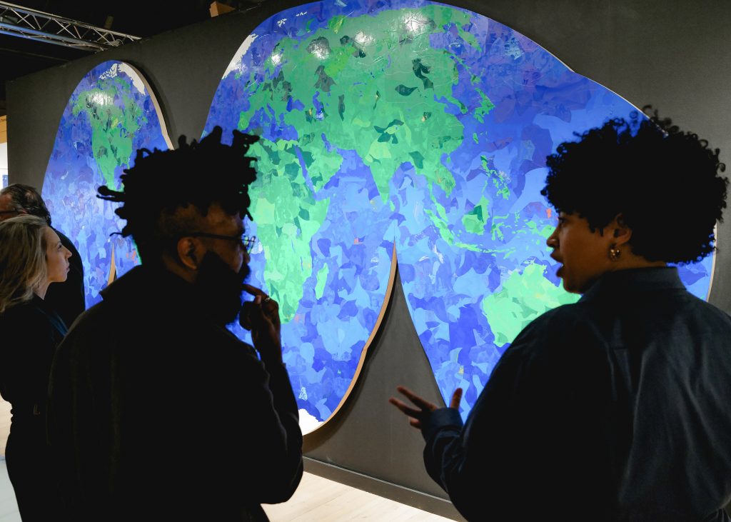 Two fairgoers stand in front of Hank Willis Thomas’s large blue and green world map artwork at an art fair, looking and gesturing toward the piece.
