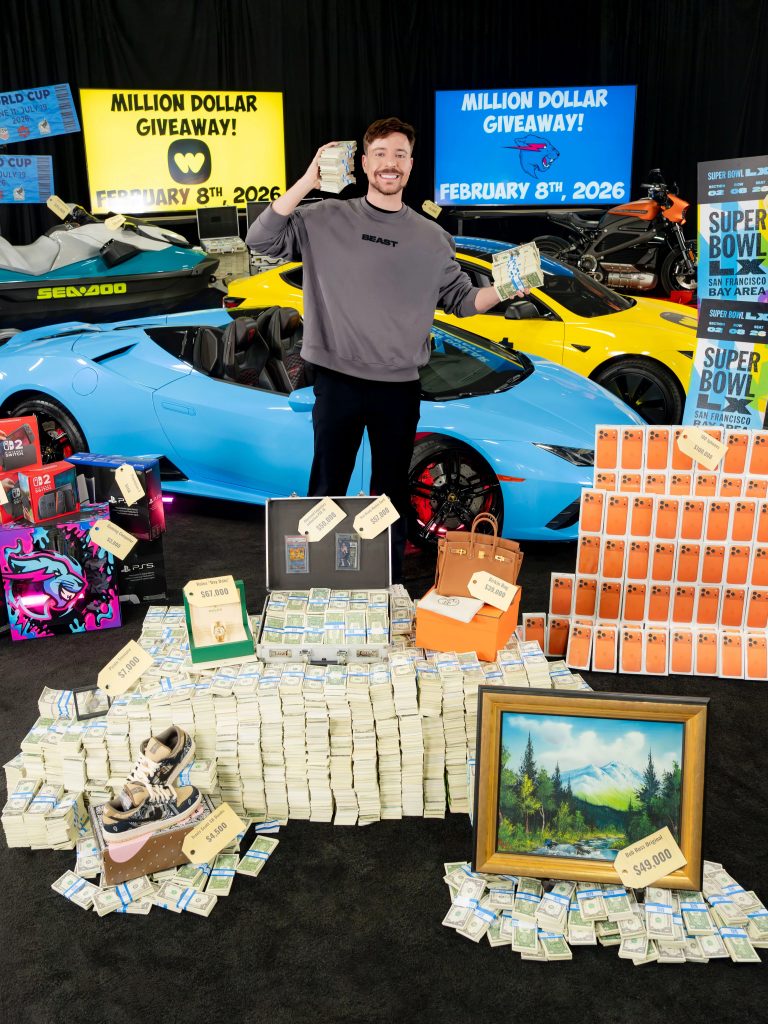 A photo of a smiling white adult man in his 20s or 30s with short brown hair and light facial hair, standing in a studio setting and holding stacks of cash in both hands. He wears a gray sweatshirt and black pants and poses in front of luxury items arranged on the floor, including large piles of bundled money, boxed electronics, designer handbags, sneakers, and a framed landscape painting. Behind him are brightly colored sports cars, a jet ski, a motorcycle, and large signs reading “Million Dollar Giveaway” with a date displayed, creating a high-energy promotional scene.