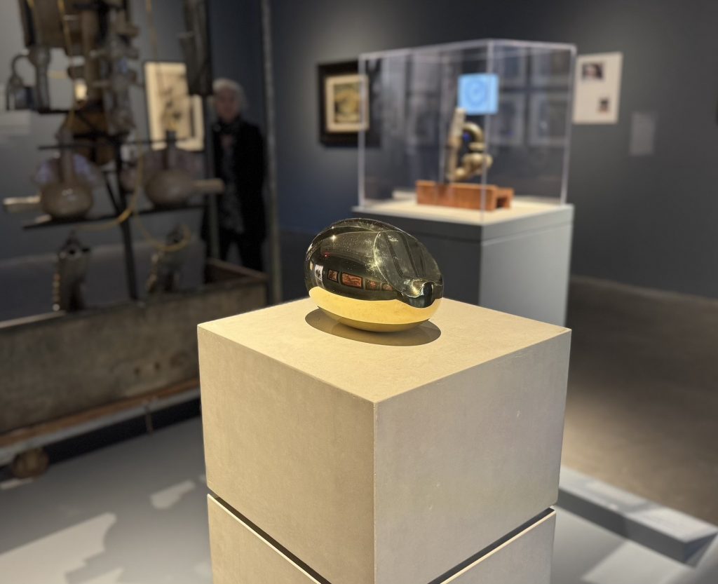 Polished, golden ovoid sculpture by Constantin Brâncuși rests on a stone pedestal, reflecting the surrounding gray gallery space and other distant modernist art installations.