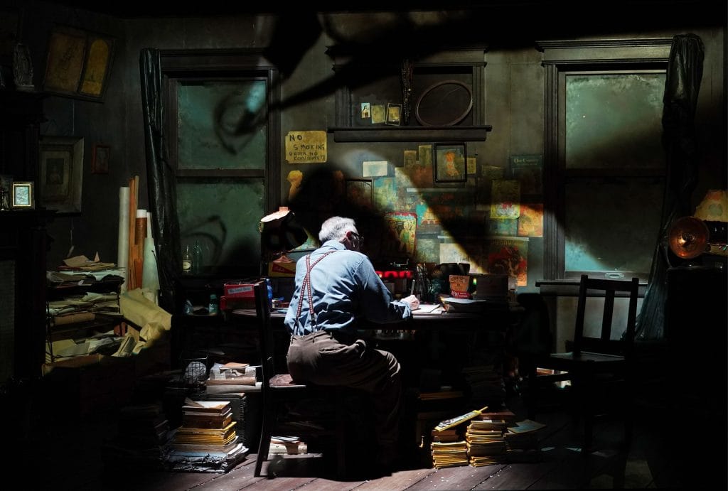 John Kelly at Henry Darger writing at cluttered desk under dramatic lighting inside dim, vintage study.