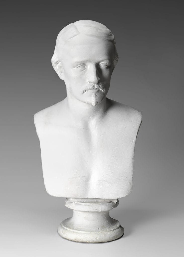 A photo of a white marble bust of a young adult white man with short parted hair and a small mustache and goatee, his gaze turned slightly to the side.