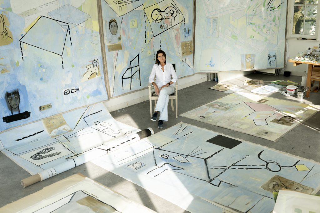 An artist sits on a chair in a spacious, sunlit studio surrounded by large-scale paintings and works on paper. The artworks, many in soft blue tones, cover the walls and floor and feature abstract lines, geometric shapes, and small figurative sketches. Several pieces are unrolled or spread out across the floor, while others hang behind the artist. Paint supplies and materials are visible on a table to the side, emphasizing an active, in-progress creative workspace.
