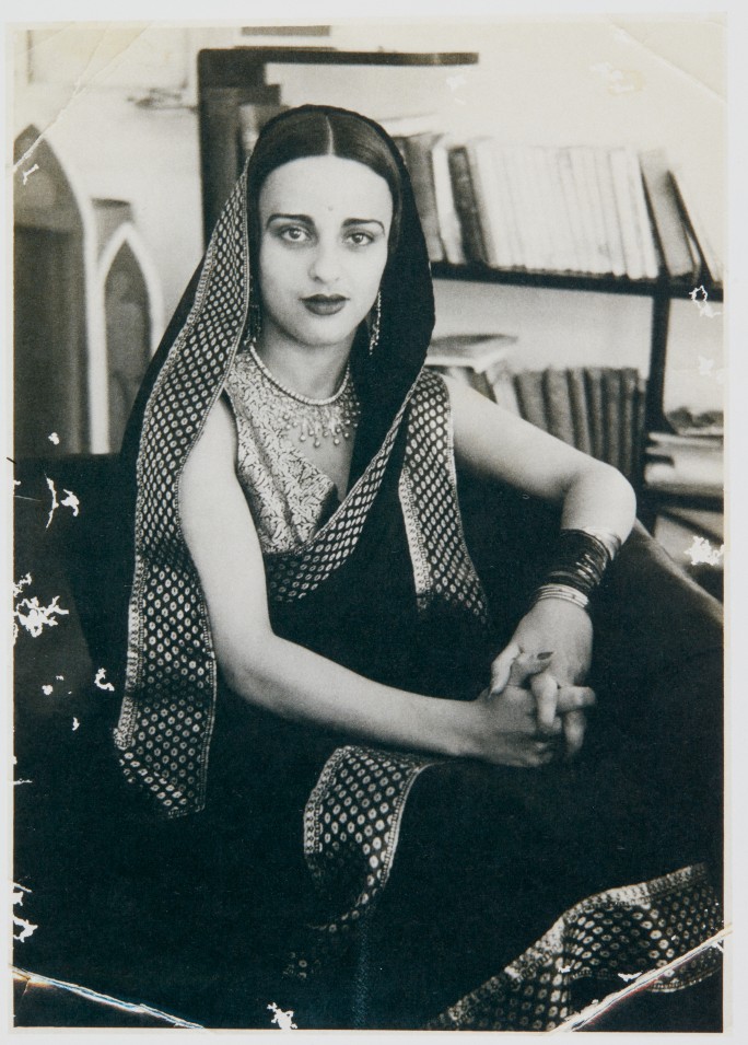 Vintage black-and-white portrait of Amrita Sher-Gil, the renowned Indian-Hungarian modernist painter, wearing a traditional sari with intricate patterns, seated in an interior with bookshelves in the background.
