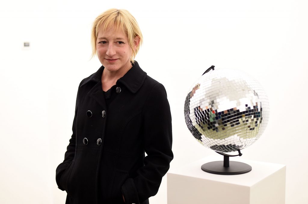 A woman in a black coat stands in a white gallery beside a mirrored disco-ball sculpture on a pedestal. She has short blond hair and a faint smile.