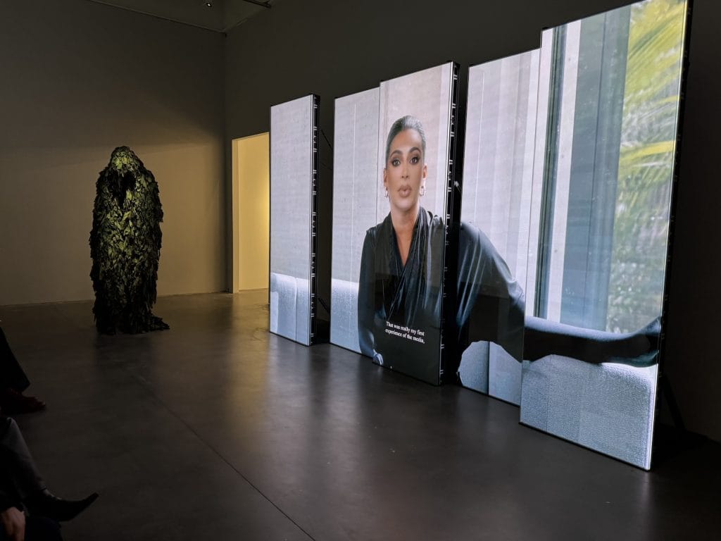 A multi-screen video installation featuring a woman speaking stands in a dark gallery, while a tall, foliage-covered ghillie suit sculpture looms in the background.
