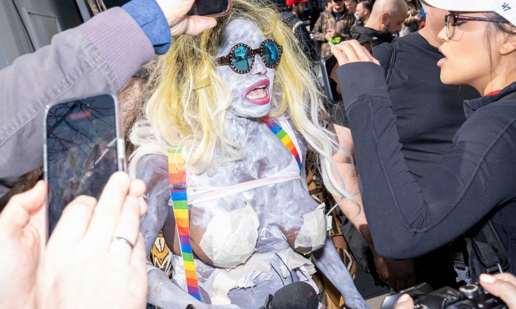 A closeup photograph of Crackhead Barney with her bare body painted white wearing just rainbow suspenders, round sunglasses, and a blonde wig in a crowd full of people