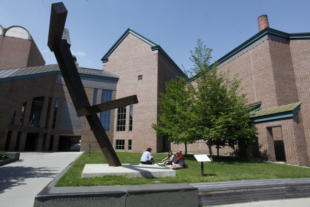 A photo of a large abstract black metal sculpture tilted at angles in a grassy plaza outside a red-brick building, with three young adults sitting on the grass nearby talking under a leafy tree on a sunny day.