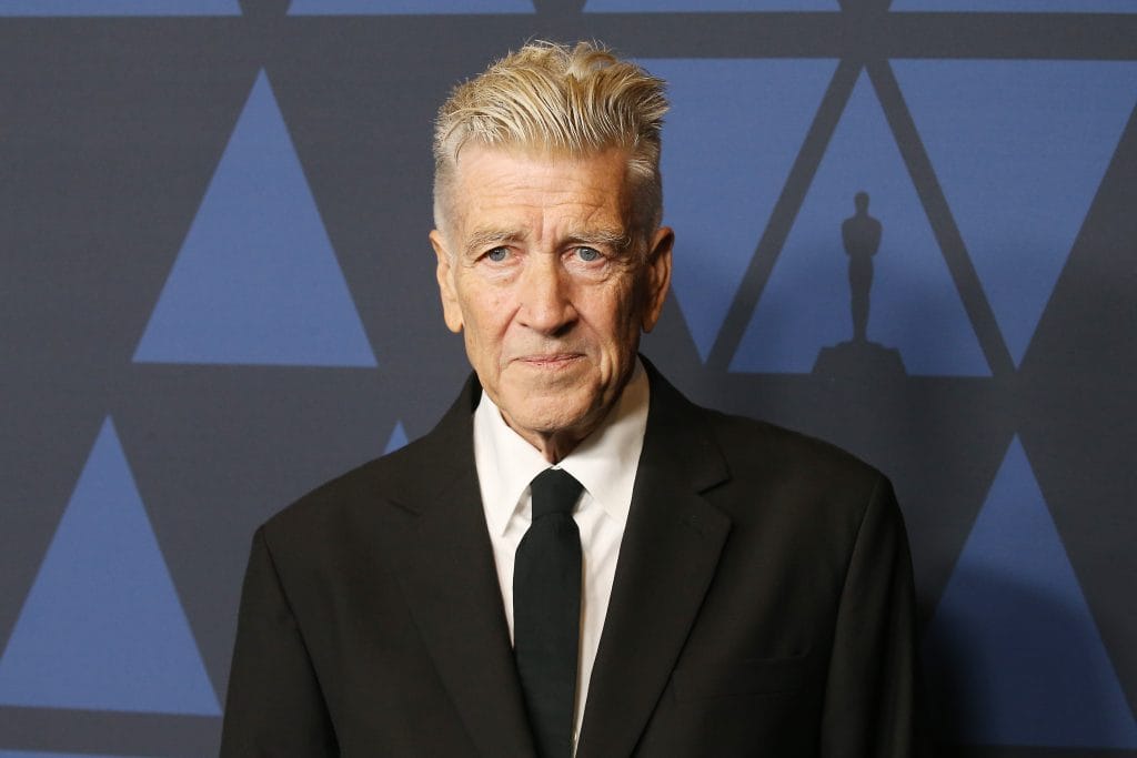 David Lynch with swept-back silver hair in black suit poses before geometric blue backdrop.
