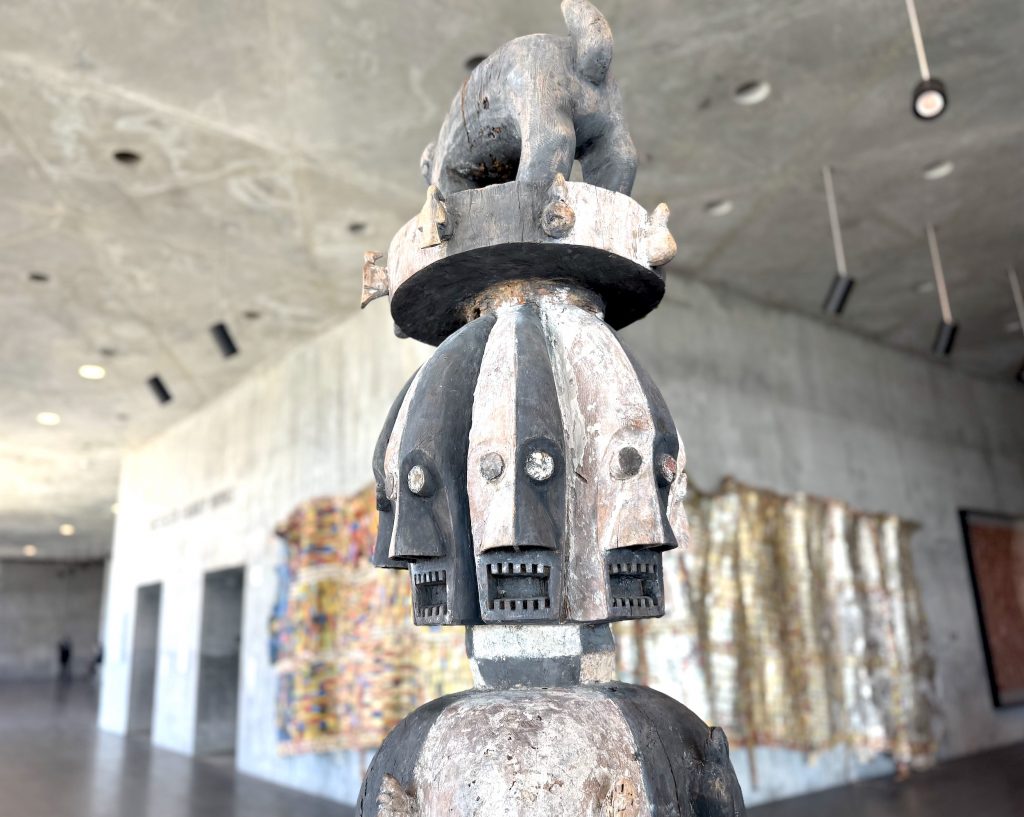 A 19th-century Ijo forest spirit figure features multiple carved faces topped by a small animal, displayed in a concrete museum gallery with vibrant wall tapestries.