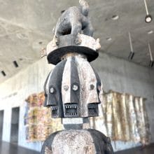 A 19th-century Ijo forest spirit figure features multiple carved faces topped by a small animal, displayed in a concrete museum gallery with vibrant wall tapestries.