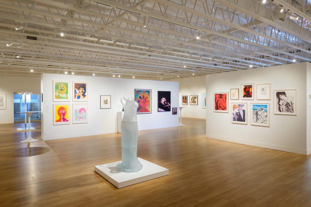 A gallery space displaying a range of colorful portrait artworks on the walls, with a sculptural dress form centered in the room.