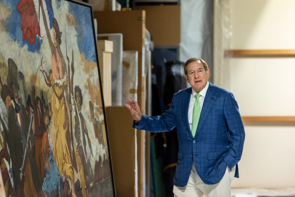 Jordan Schnitzer stands beside a large painting in a warehouse, gesturing toward the artwork during a guided tour.