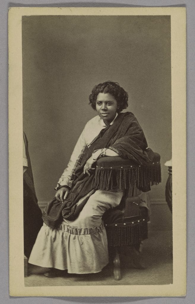 A black and white photo of a young Black woman, likely in her 20s or 30s, with dark curly hair parted at the center, seated and leaning on a fringed chair, wearing a shawl and dress, looking directly at the camera with a composed expression.