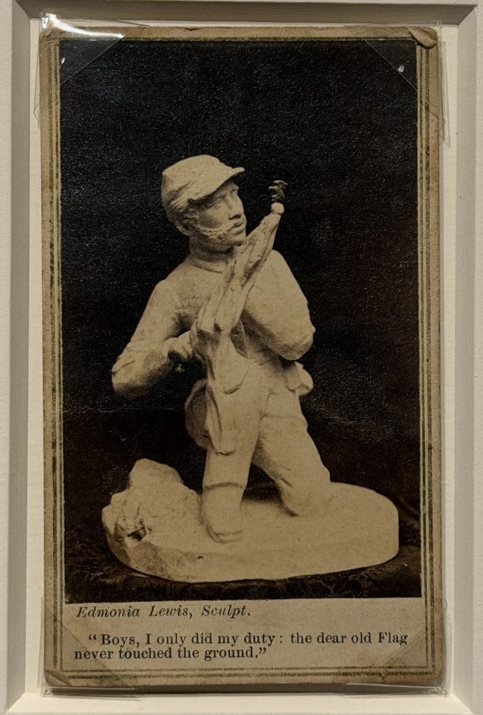 A photo of a framed vintage print showing a sculpted young Black Civil War soldier in uniform kneeling and holding a flag close to his chest, with a caption below crediting Edmonia Lewis and including a quote about duty and the flag.