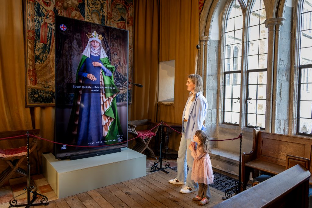 Woman and child watch interactive screen portraying Eleanor of Castile speaking inside historic chapel room.