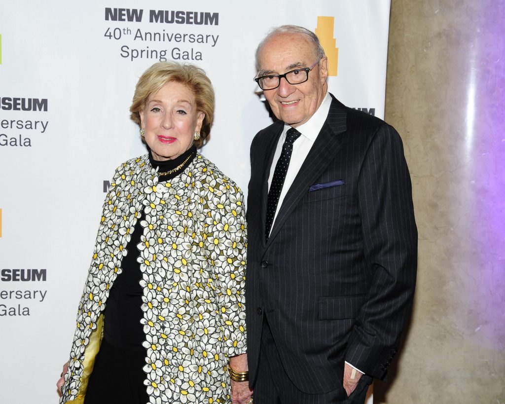 A photo of an older white couple dressed formally at a gala. The woman has short blonde hair and wears a black outfit with a long, floral-patterned coat covered in white and yellow daisies. The man has short gray hair and glasses and wears a dark pinstriped suit with a white shirt and black tie. They stand smiling in front of a backdrop for the New Museum’s 40th Anniversary Spring Gala.