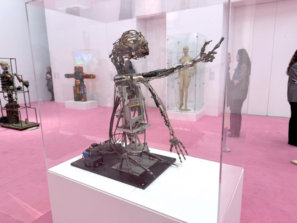 An intricate, mechanical E.T. animatronic stands in a glass case on a white pedestal, surrounded by other robotic figures in a gallery with pink floors.