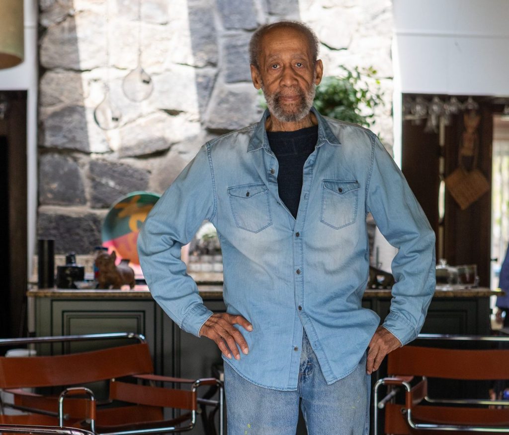 A photo of an older Black man with short gray hair and a beard standing indoors with his hands on his hips. He wears a light denim shirt over a dark shirt and light jeans. Behind him is a stone wall and a warmly lit room with modern furniture and decorative objects.